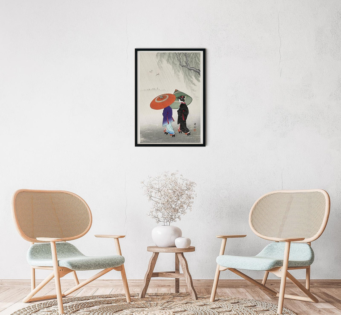 A framed poster of 'Two Women in the Rain' by Ohara Koson, displayed on a wall above a small round table with a vase, next to two modern chairs.