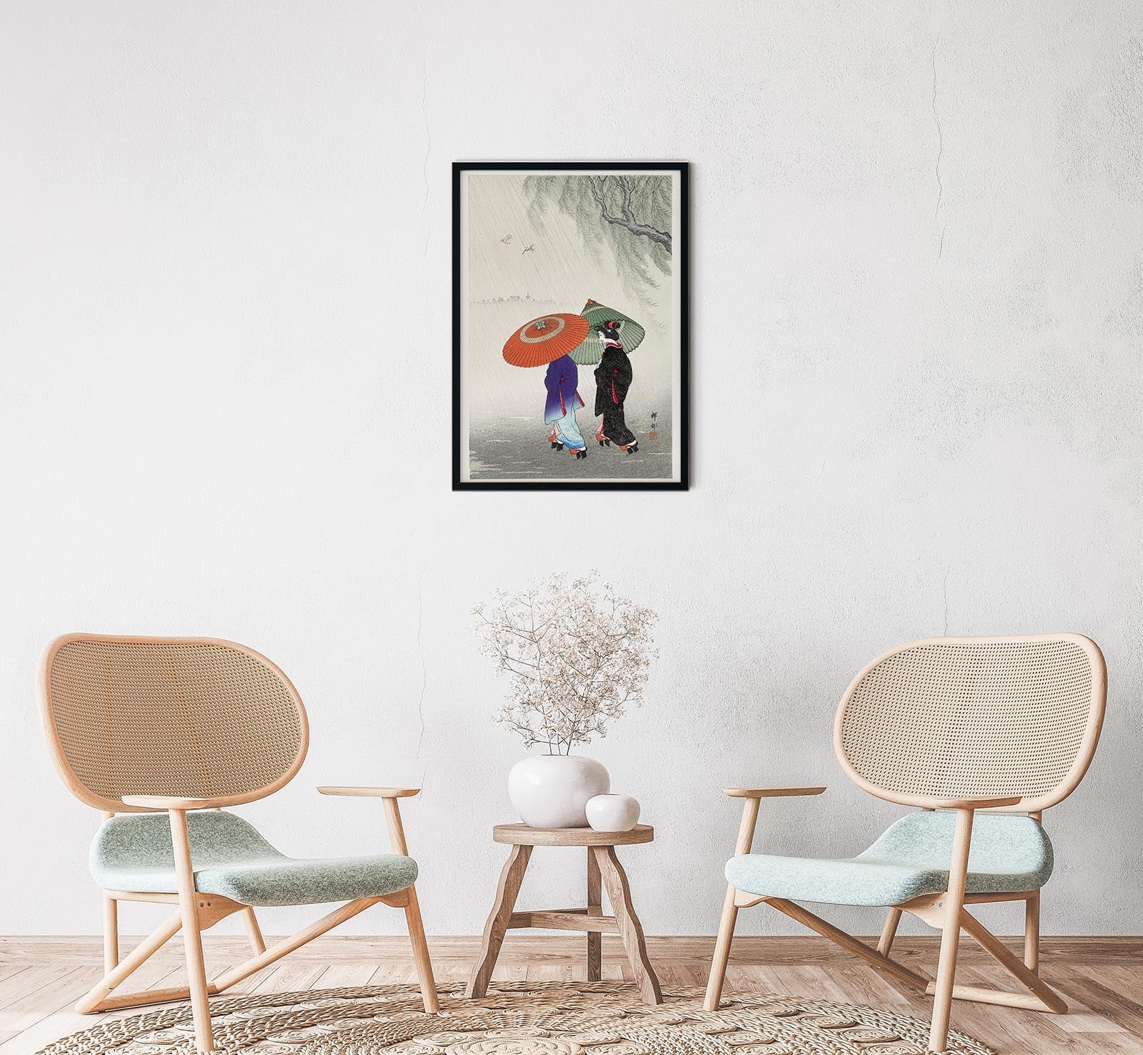 A framed poster of 'Two Women in the Rain' by Ohara Koson, displayed on a wall above a small round table with a vase, next to two modern chairs.